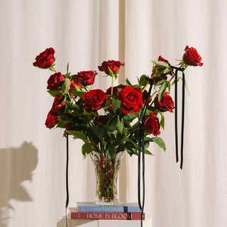 Bouquet of red roses in a clear vase with black ribbons against a white curtain.